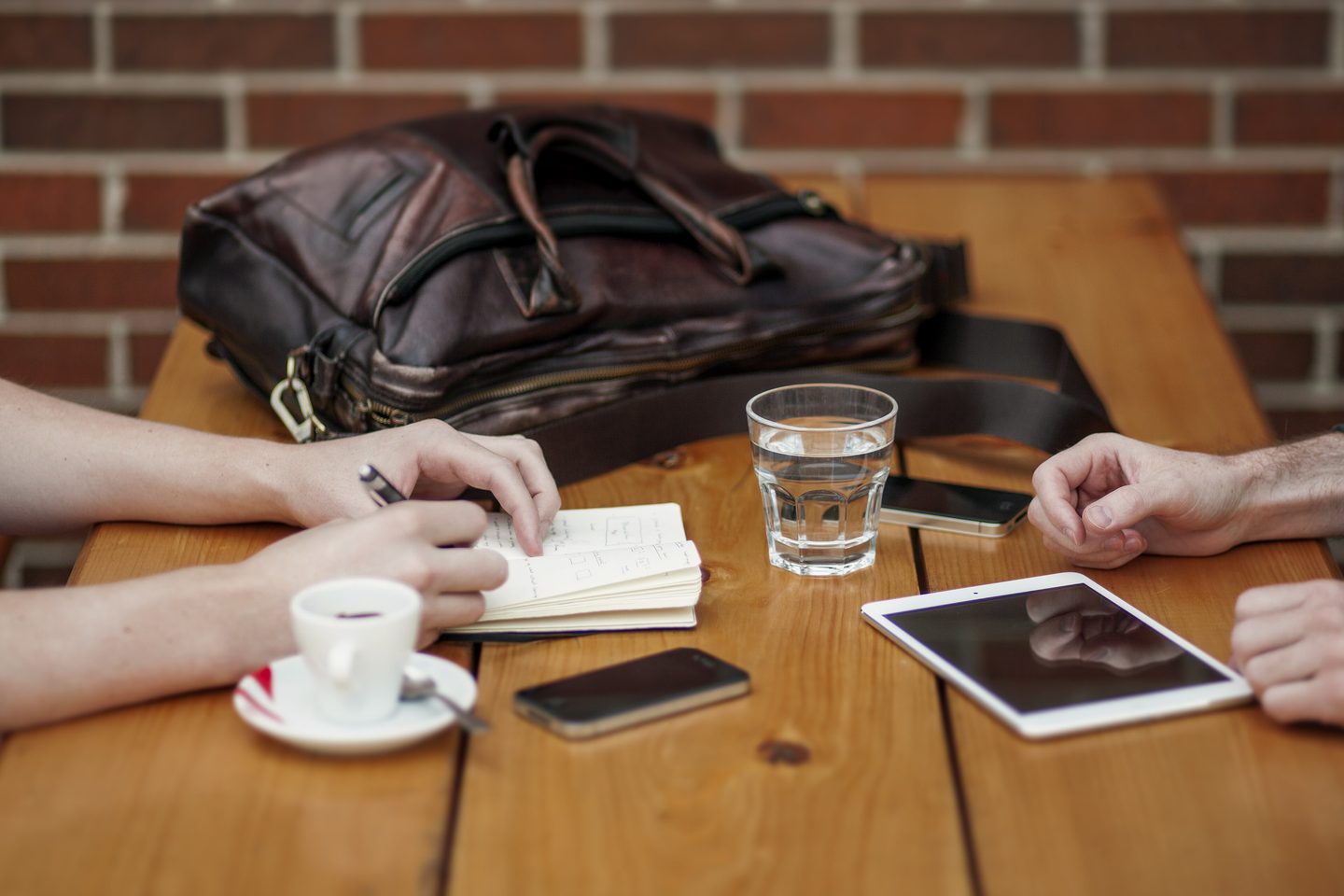 Duas pessoas conversando em uma mesa de madeira, com caderno de anotações, tablet, celulares e café.